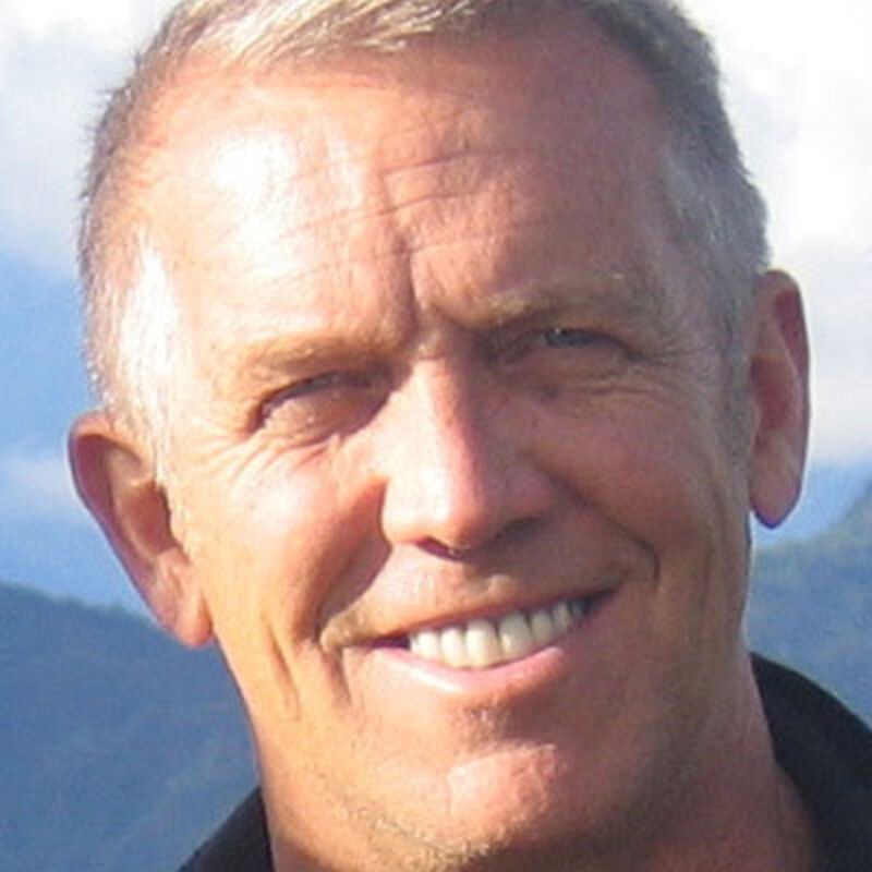 The image shows a close-up of a middle-aged man with short, graying hair and a warm smile. He has fair skin and some wrinkles around his eyes, suggesting age and experience. The background features a blurred landscape with mountains and a blue sky, indicating the photo was taken outdoors. The man is wearing a dark shirt, which contrasts with the brighter background.
